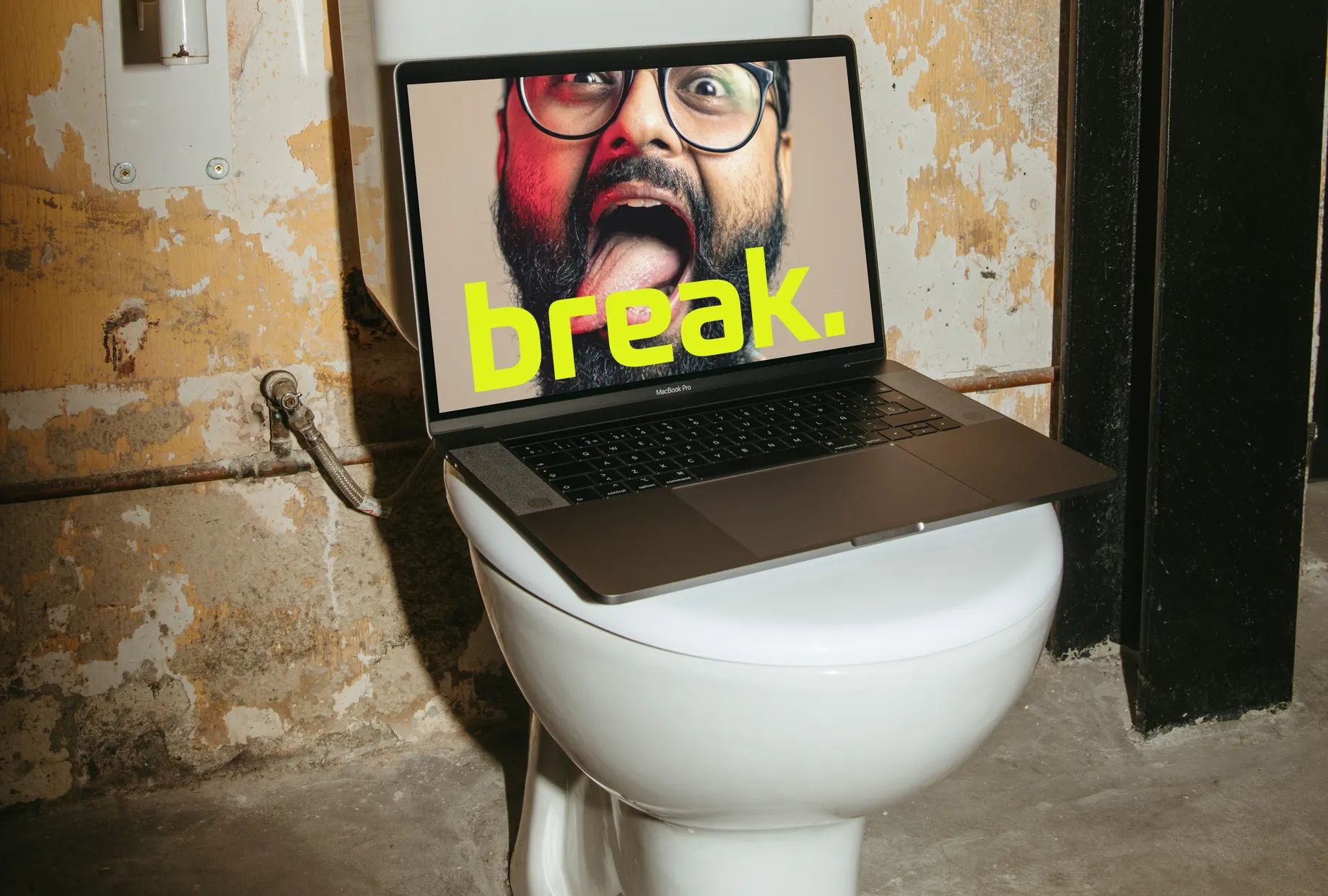 Laptop auf einer Toilettenschüssel in einem Rohbau-WC. Auf dem Bildschirm ein Mann mit Brille, der die Zunge herausstreckt, darüber der Schriftzug „break.“ in Neongelb.