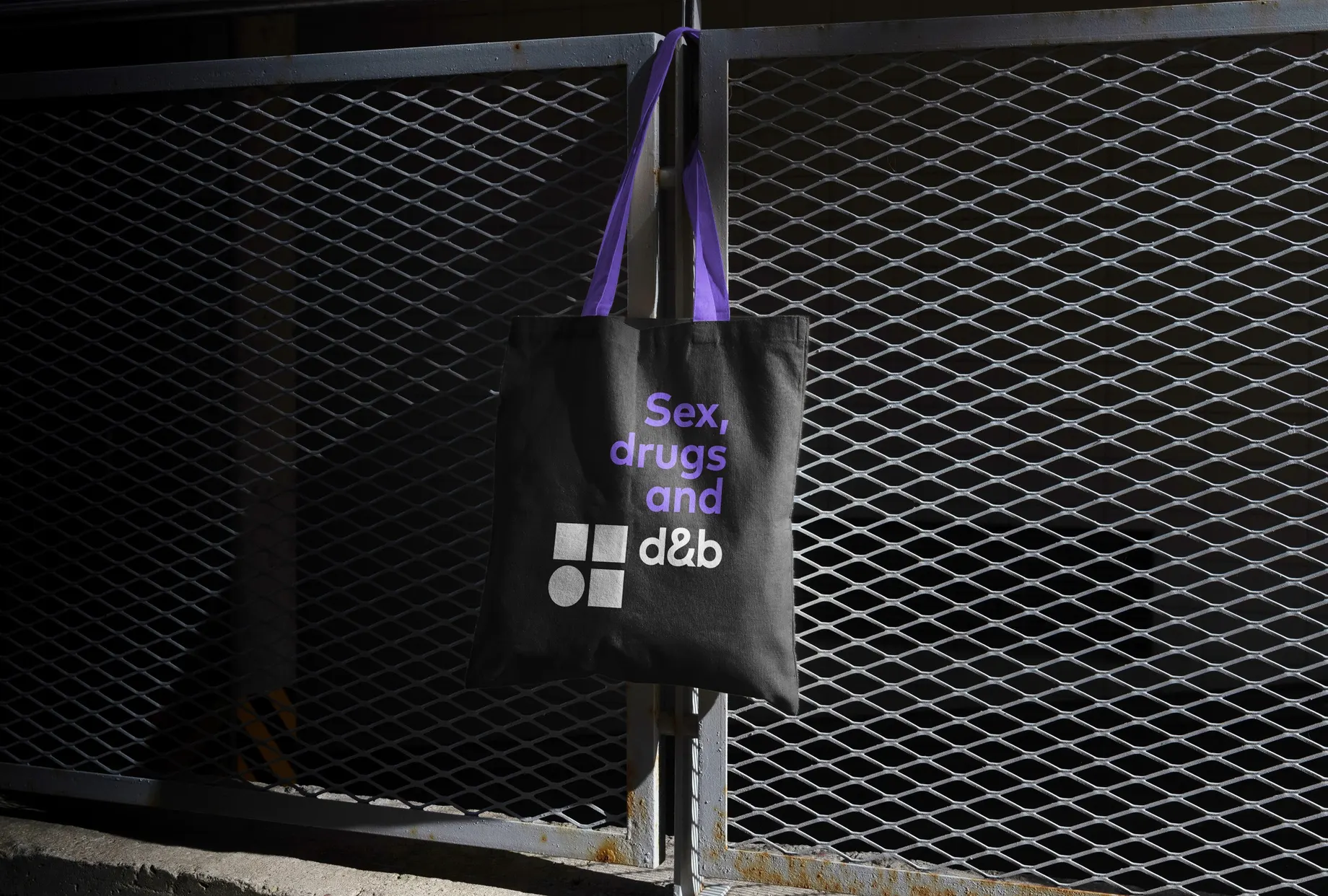 Schwarze Stofftasche mit violetten Henkeln, an einen Metallzaun gehängt. Aufdruck: „Sex, drugs and“ über dem d&b-Logo.