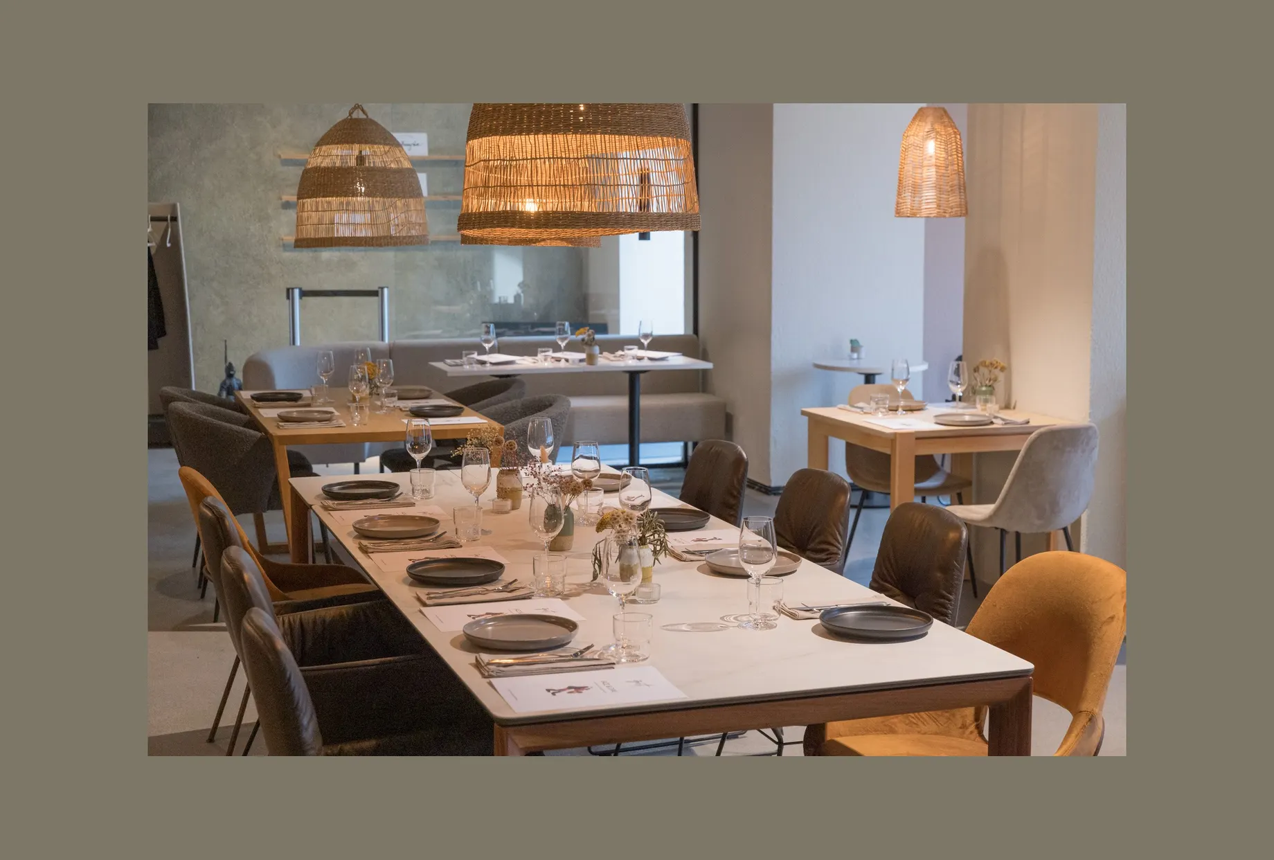 Gastraum mit langer gedeckter Tafel: helle Tischplatte, dunkle Teller, Weingläser und kleine Blumenvasen. Darüber grosse Korbleuchten, im Hintergrund weitere Tische und eine Sitzbank.