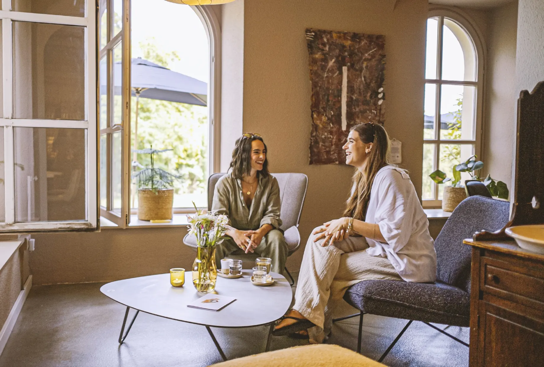 Zwei Frauen sitzen lachend in Sesseln um einen ovalen Couchtisch, darauf eine Vase mit Lilien, Kerzen und Getränke. Durch das geöffnete Fenster fällt Licht in den Raum.