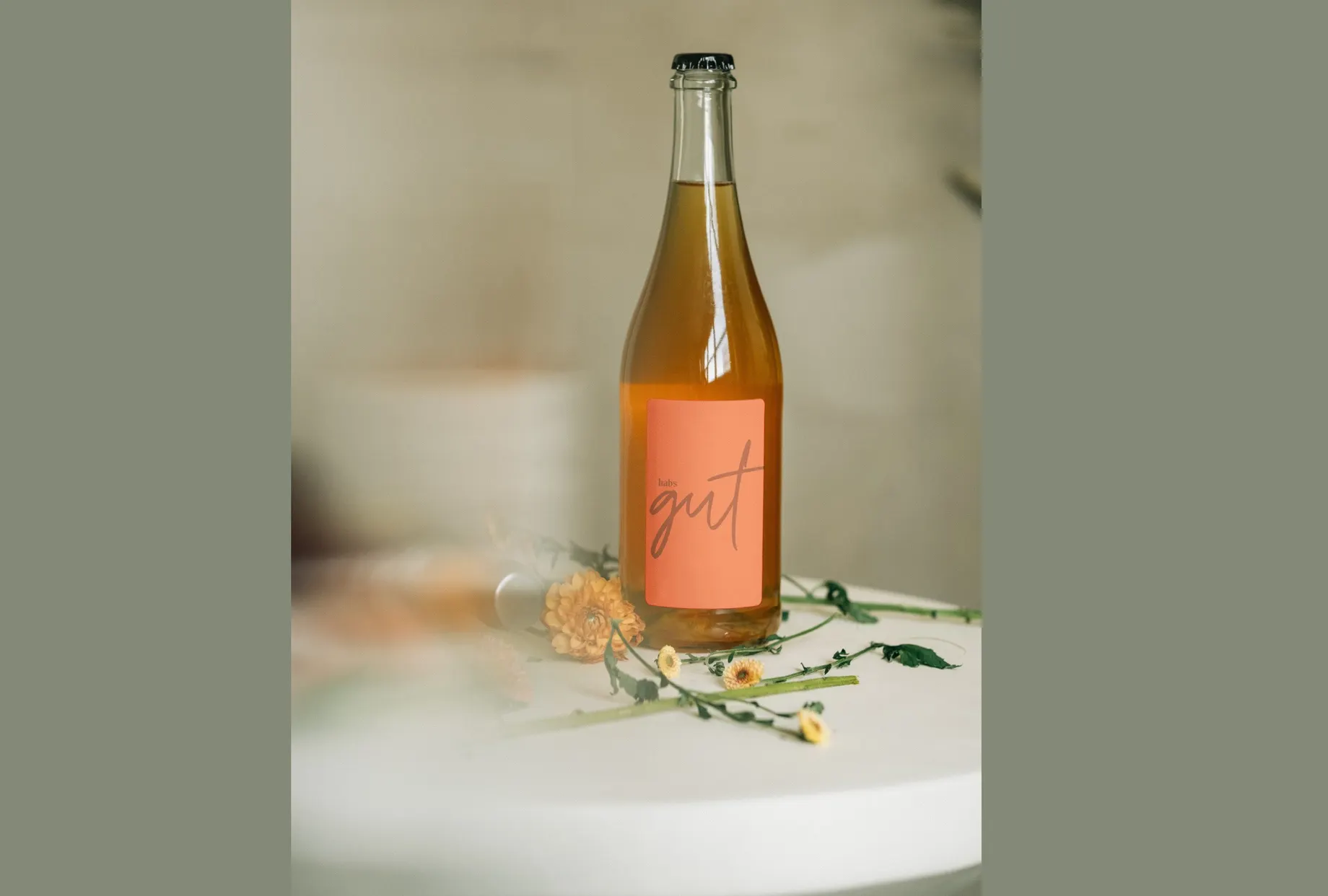 Bügelflasche mit hellem, trübem Getränk auf einem runden weissen Tisch. Das lachsfarbene Etikett trägt den habs*gut-Schriftzug, daneben liegen getrocknete Blüten.