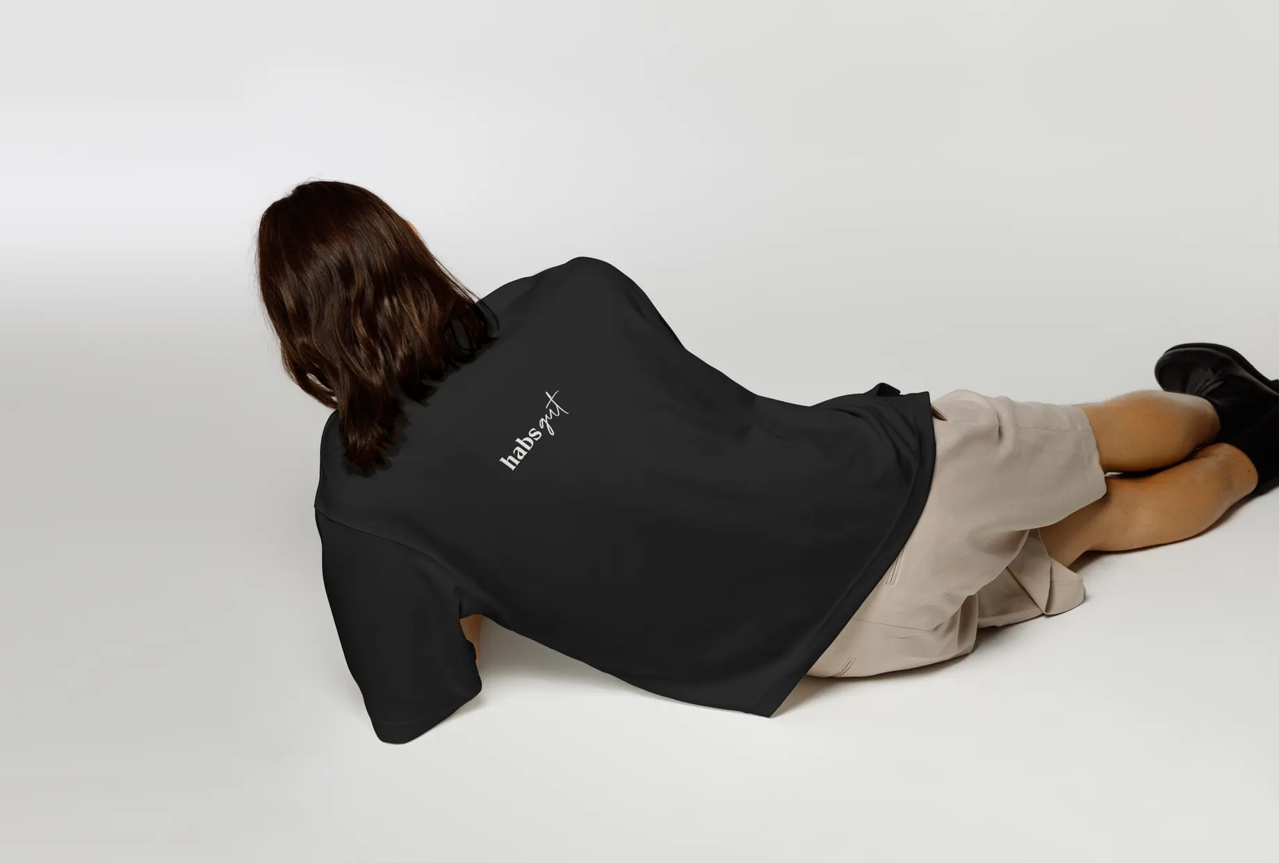 Person in schwarzem Oversize-Shirt liegt auf hellem Studiogrund, das Gesicht vom Haar verdeckt. Auf dem Rücken der habs*gut-Schriftzug in Weiss.