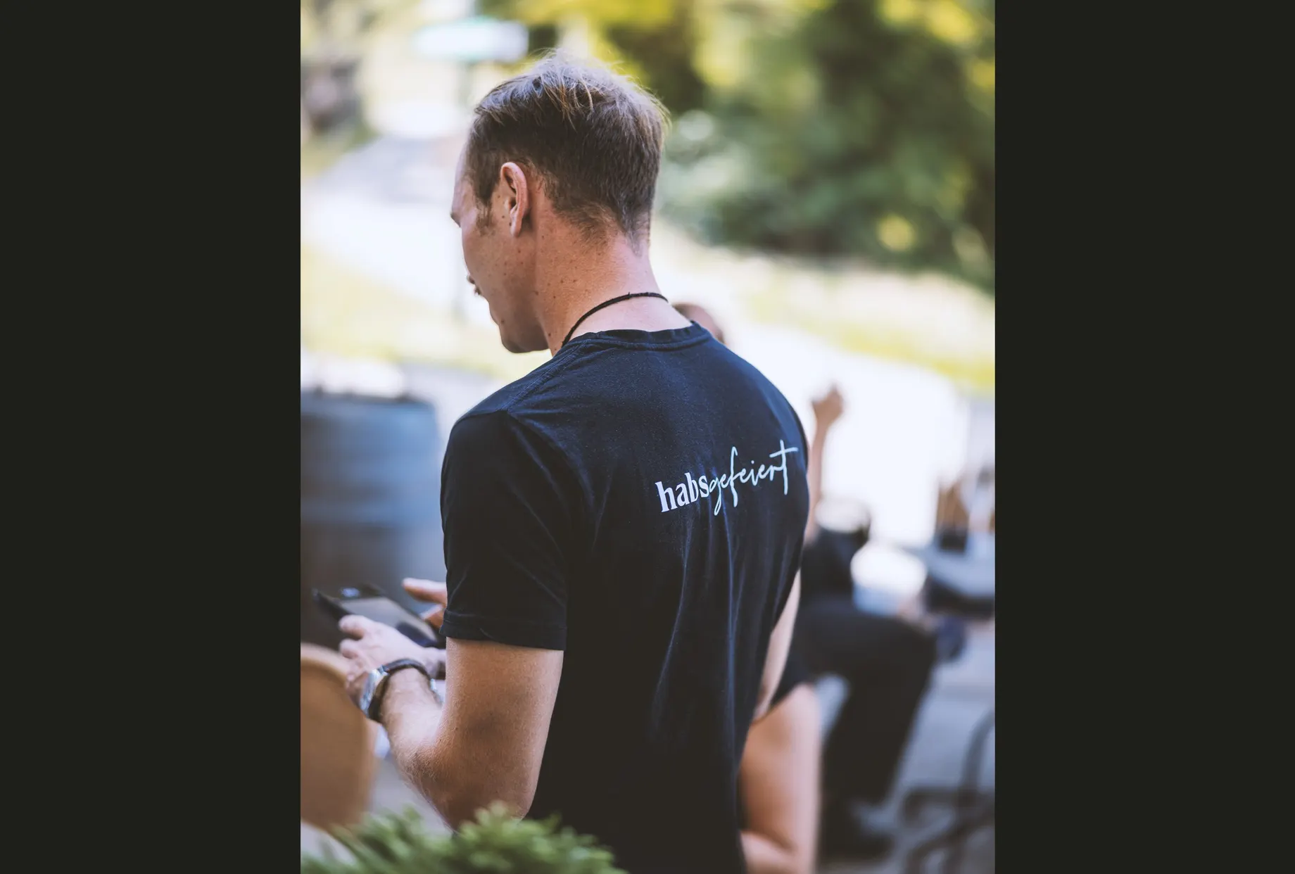 Person von hinten in schwarzem T-Shirt mit dem Aufdruck „habsgefeiert“ auf dem Rücken, im Freien vor unscharfem Grün.