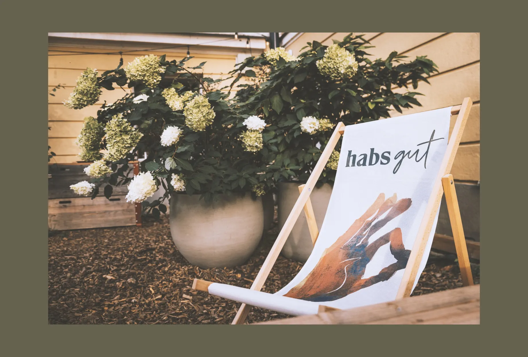 Liegestuhl mit dem habs*gut-Schriftzug auf der Bespannung, im Garten zwischen blühenden Hortensien.