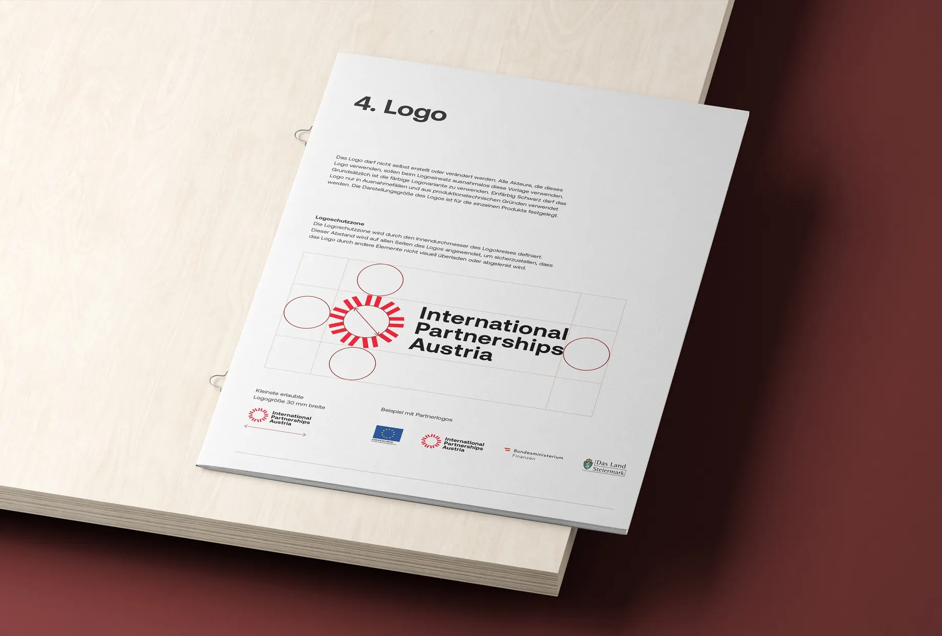 Aufgeschlagener Guide auf hellem Holz, Kapitel „4. Logo“: Regeln zur Logoschutzzone mit eingezeichnetem Raster, dazu die kleinste erlaubte Grösse und ein Beispiel mit Partnerlogos.