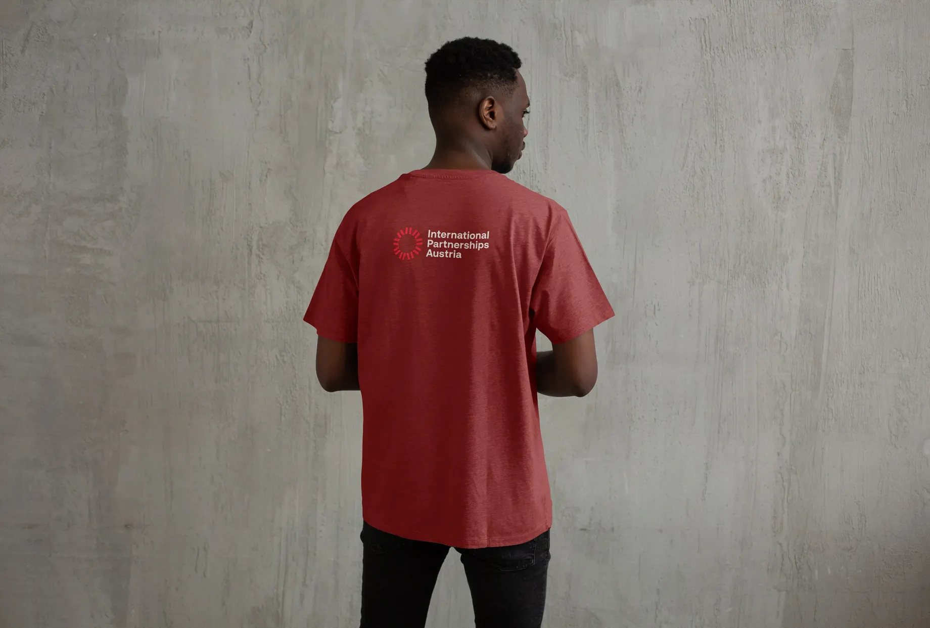 Person von hinten vor einer Betonwand, in rotmeliertem T-Shirt mit dem Logo „International Partnerships Austria“ zwischen den Schulterblättern.