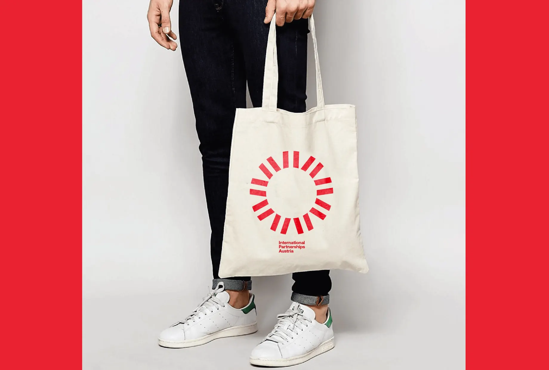 Person trägt eine helle Baumwolltasche mit dem grossen roten Strahlenkreis und dem Schriftzug „International Partnerships Austria“, seitlich gerahmt von roten Flächen.