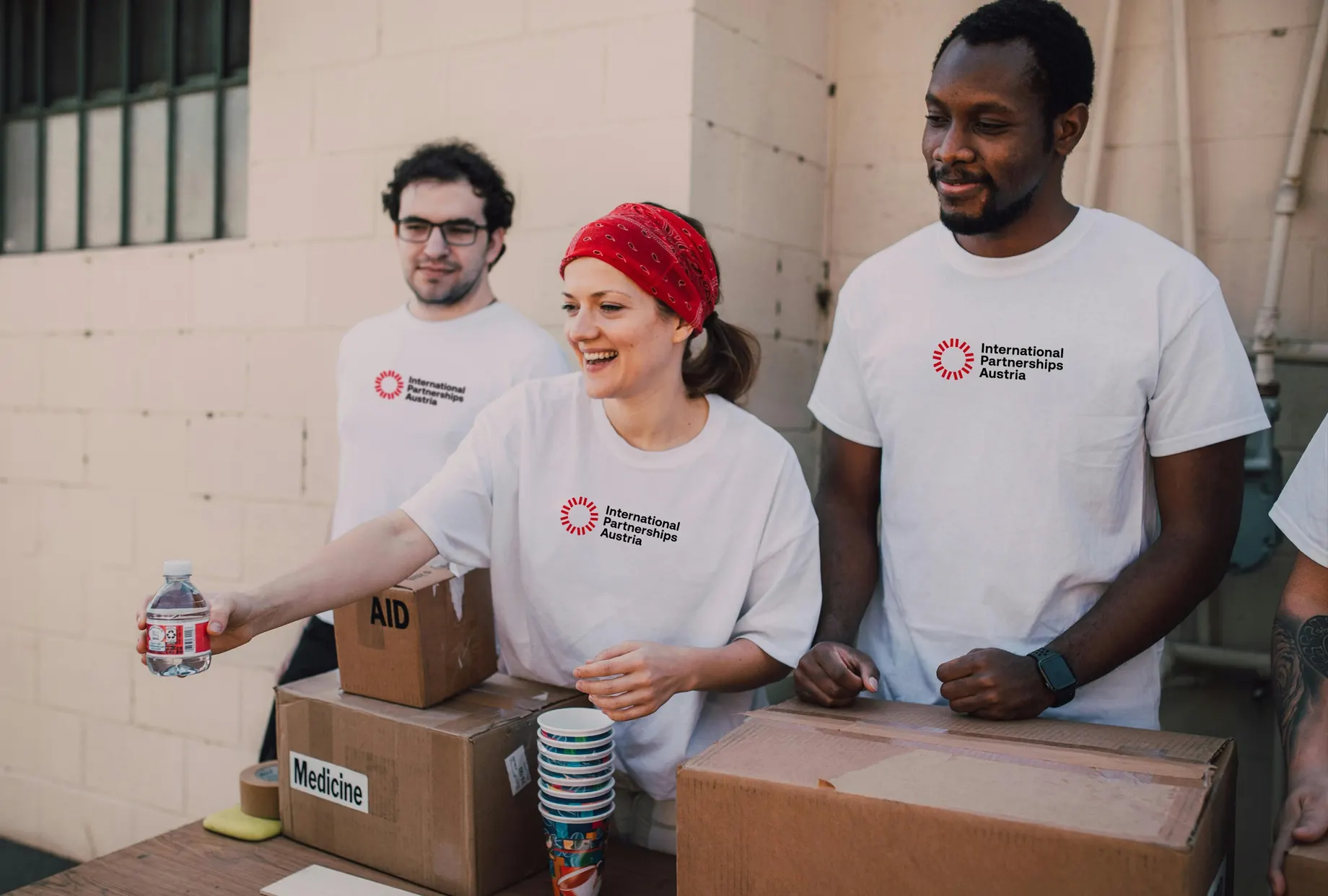 Drei Personen in weissen T-Shirts mit dem Logo „International Partnerships Austria“ stehen hinter Kartons mit den Aufschriften „AID“ und „Medicine“ und reichen eine Wasserflasche weiter.