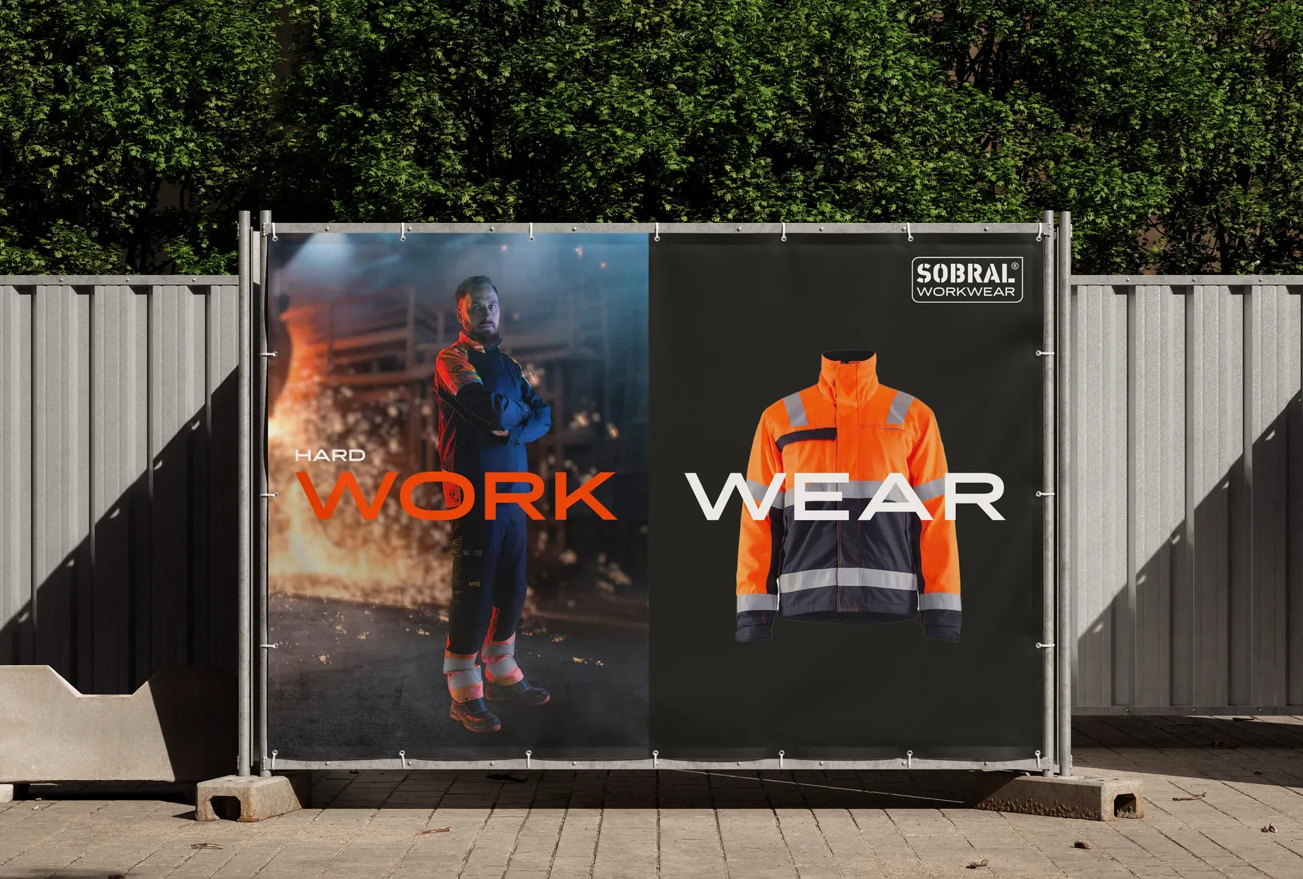 Bauzaunplane vor einer Hecke: links ein Mann in Arbeitskleidung vor Funkenflug mit der Zeile „Hard work“, rechts eine orange Warnschutzjacke vor Schwarz mit dem Wort „Wear“.