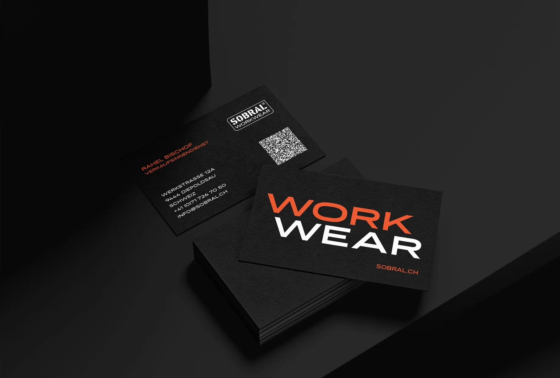 Schwarze Visitenkarten auf dunklem Grund: hinten die Rückseite mit Kontaktdaten und QR-Code, vorne ein Stapel mit dem Schriftzug „Workwear“ in Orange und Weiss.