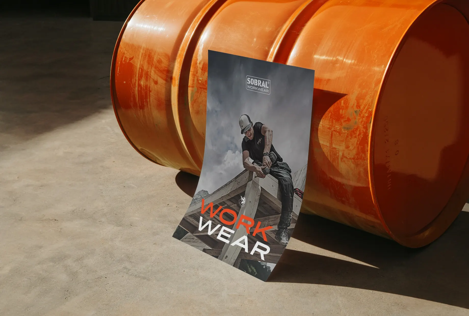 Plakat an ein orangefarbenes Ölfass gelehnt: ein Zimmerer mit Kappe arbeitet auf einem Holzdachstuhl, darüber der Schriftzug „Work Wear“.