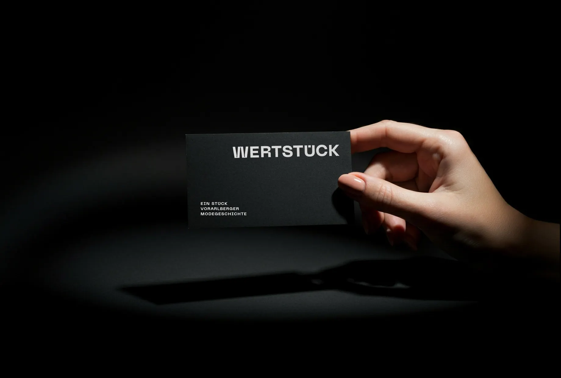 Eine Hand hält eine schwarze Visitenkarte ins Spotlicht. Aufdruck: „Wertstück – ein Stück Vorarlberger Modegeschichte“.