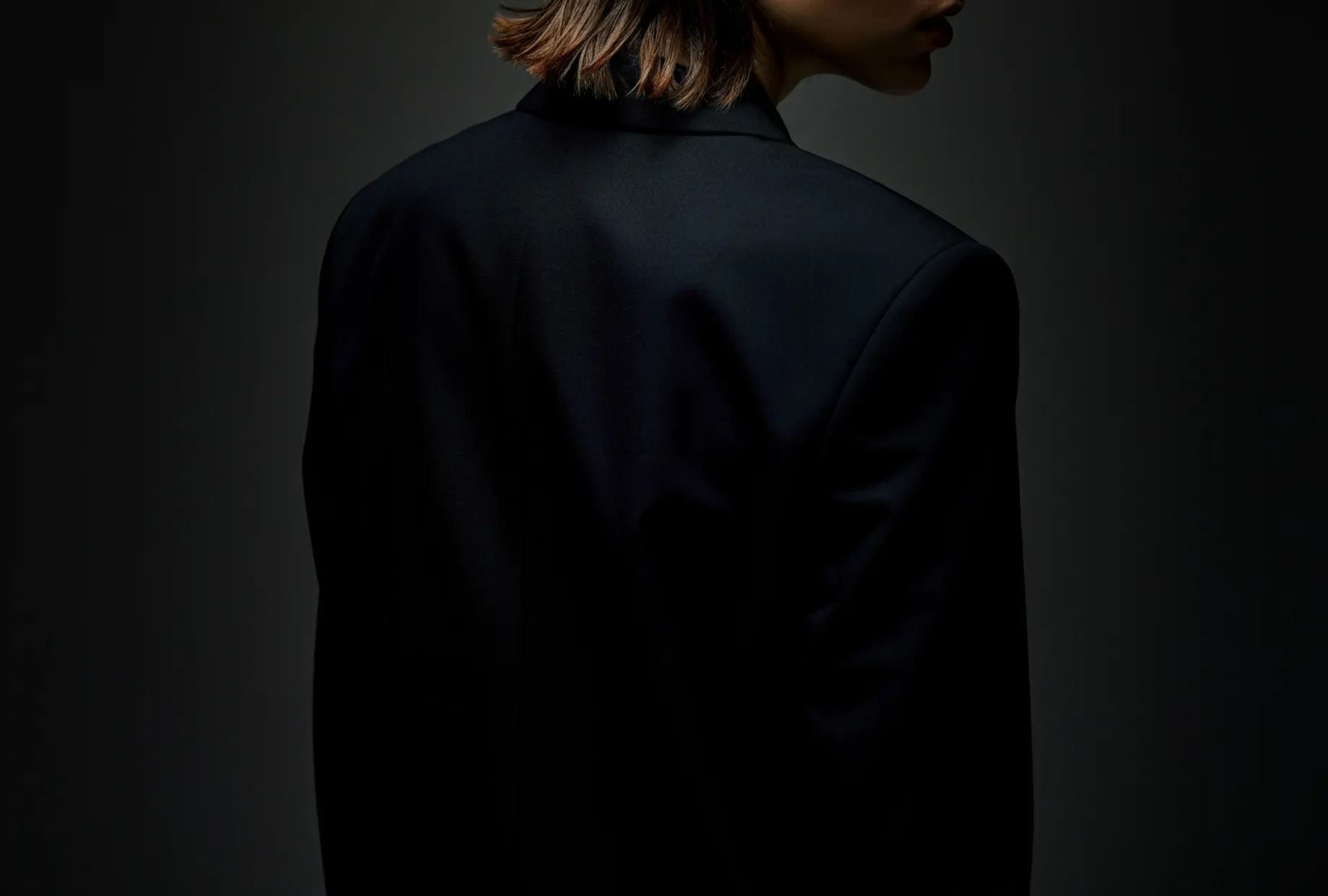 Person von hinten in einem dunklen Blazer mit betonter Schulterpartie, vor grauem Hintergrund im Halbdunkel.